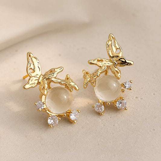 Close-up of gold butterfly earrings with textured wings, glass center, and zircon stones.