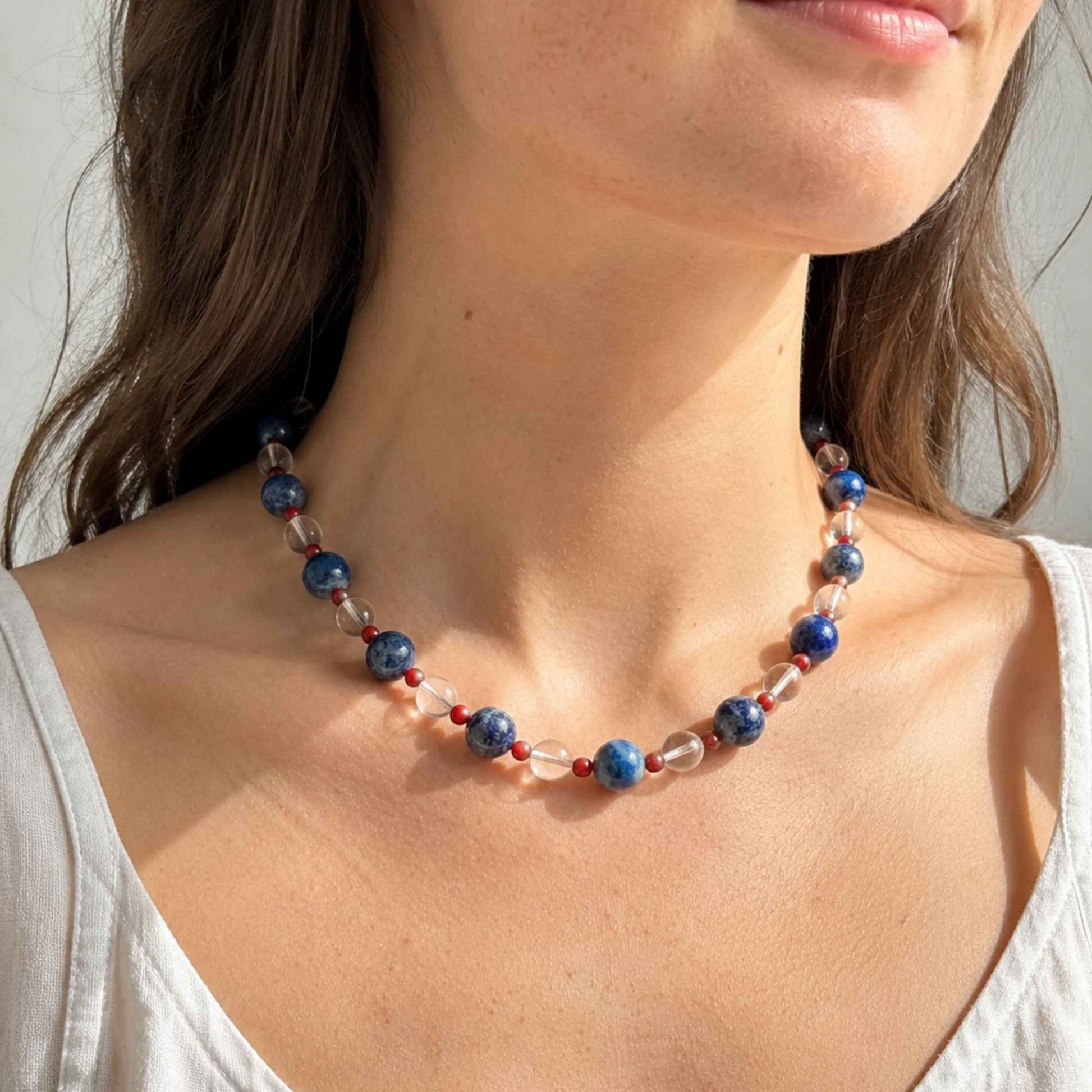 Model wearing a blue, red, and clear natural stone necklace at the collarbone in warm sunlight.
