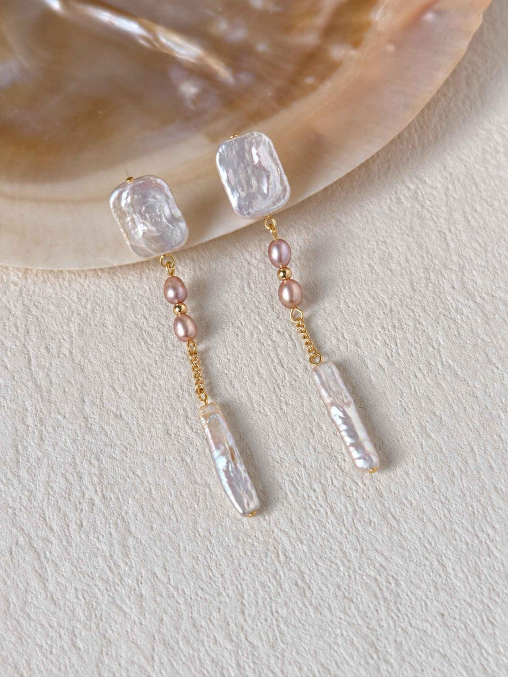 Cadence long freshwater pearl earrings styled beside a natural shell on a textured ivory surface.