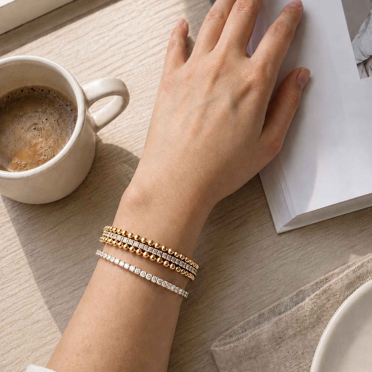 Crossline CZ Bracelet layered with gold bead and slim stone-set bracelets beside a coffee cup