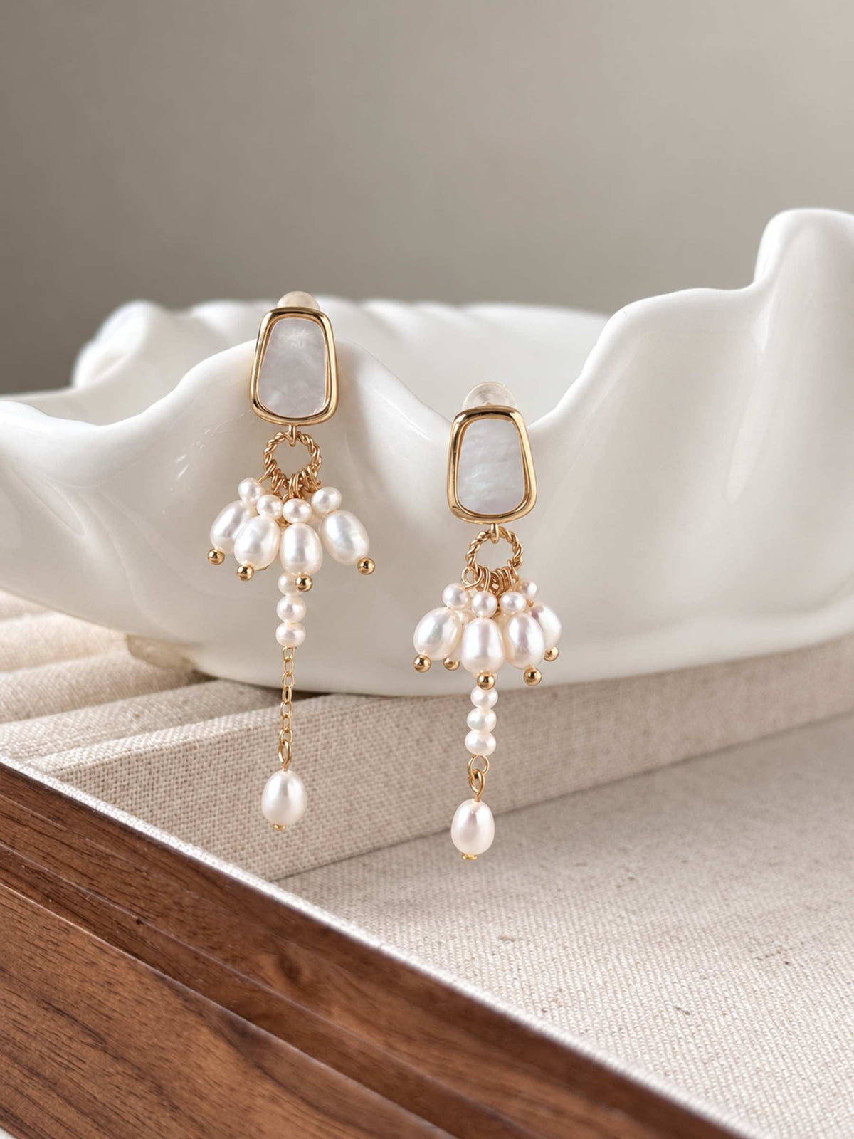 Drape asymmetric pearl earrings displayed on a white sculptural dish with delicate gold-tone details.