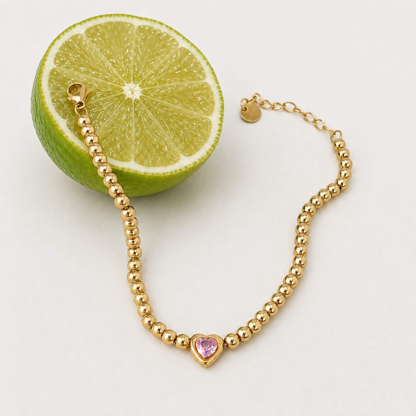 Gold bead and pink heart bracelet styled beside a fresh lime on a light surface