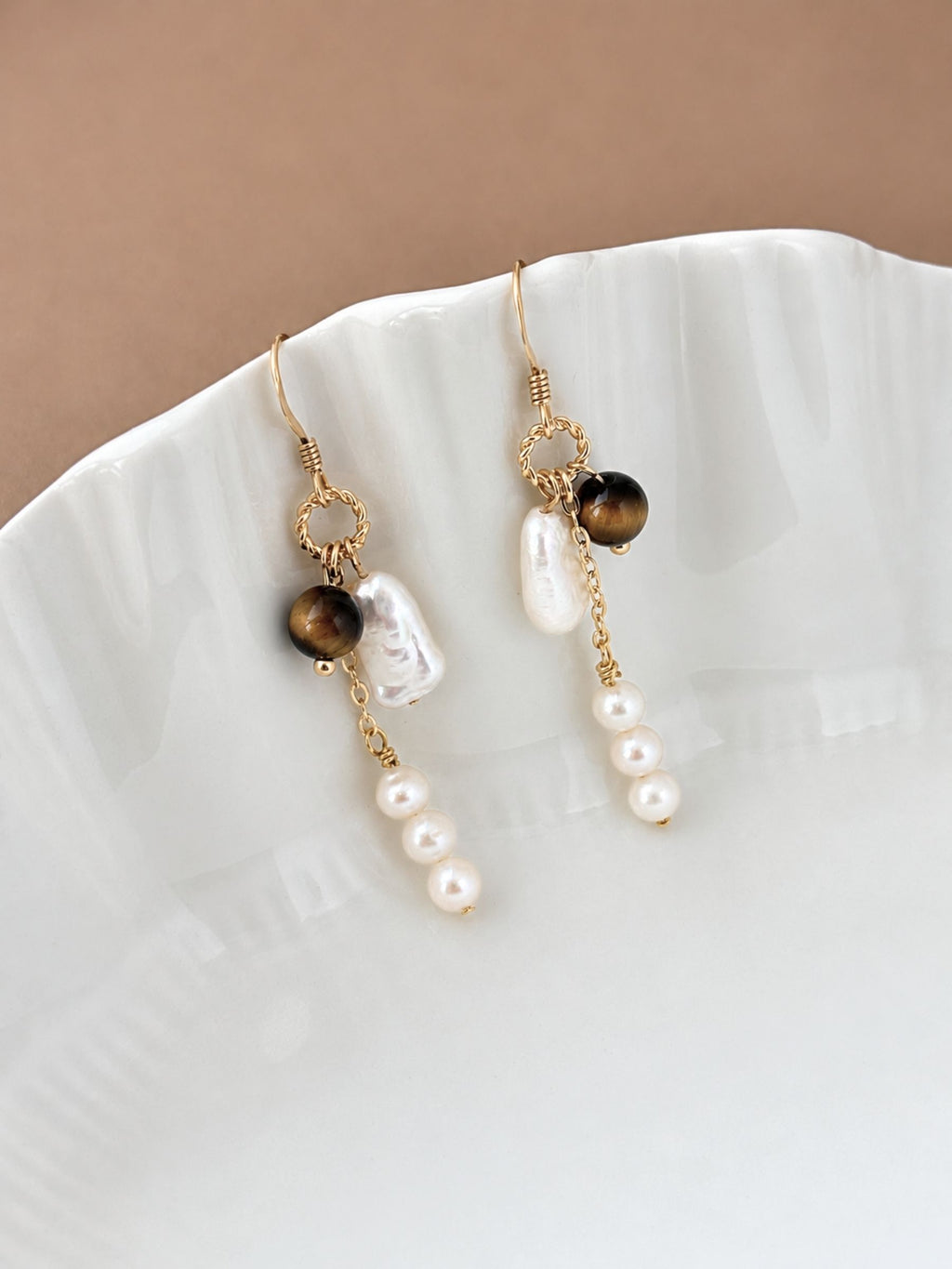 Orbit pearl and tiger eye earrings styled on a white ceramic dish, highlighting the warm brown stone and luminous pearls.
