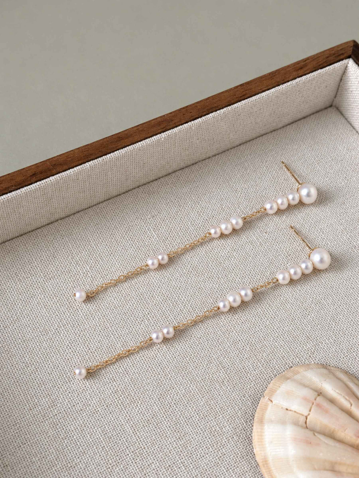 Long freshwater pearl earrings arranged on a linen jewelry tray beside a natural shell.
