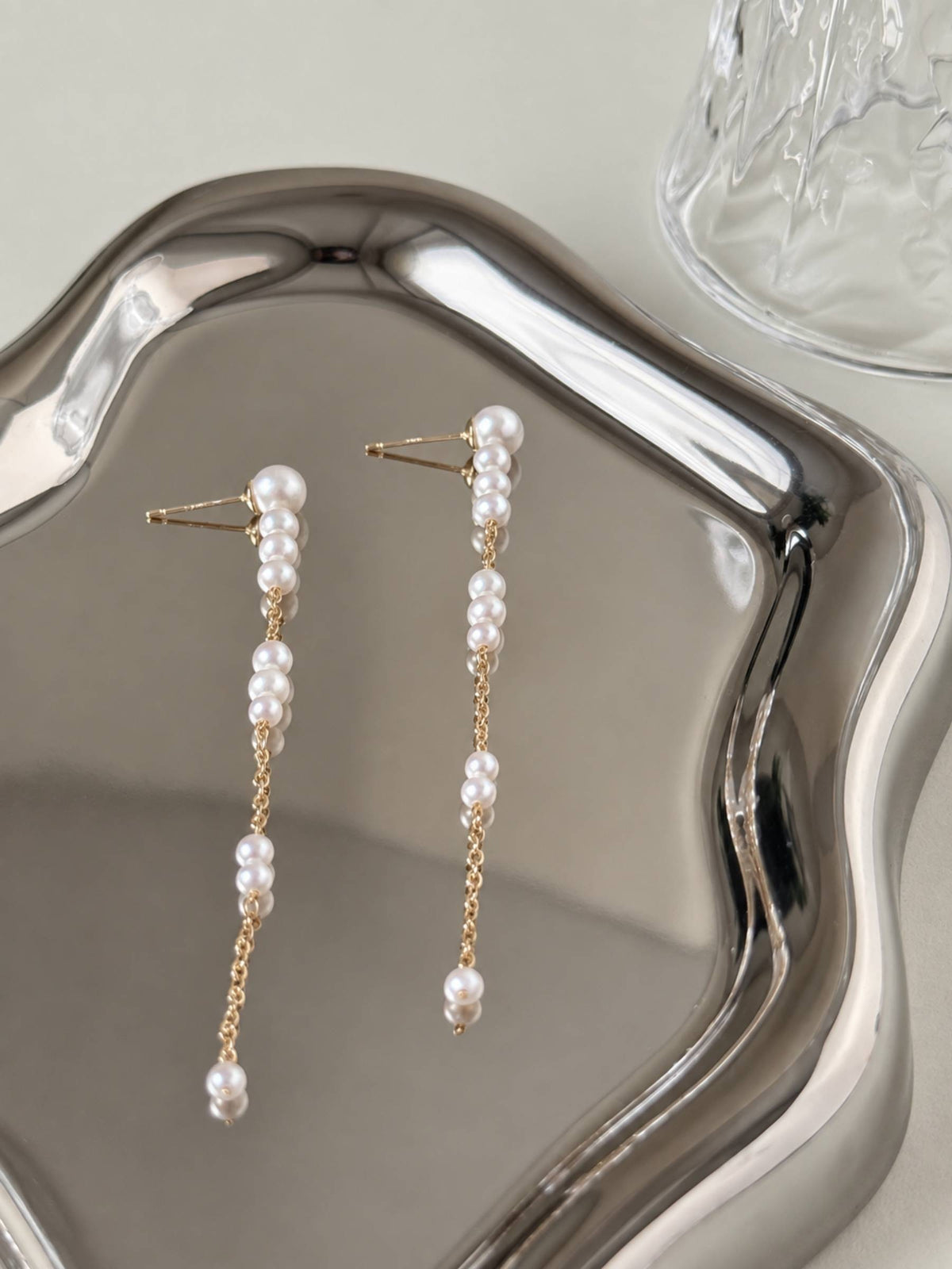 Pace pearl chain earrings arranged on a reflective silver tray, showing their long linear silhouette.