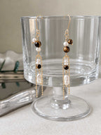 Tether tiger eye and freshwater pearl earrings hanging from a clear glass display with long gold-tone chain drops.