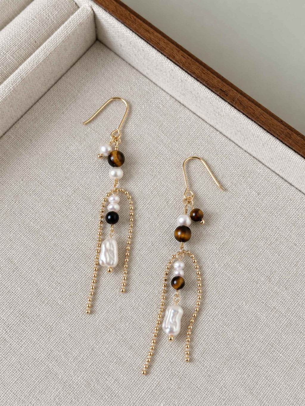Tether natural tiger eye and pearl earrings arranged on a neutral linen jewelry tray.